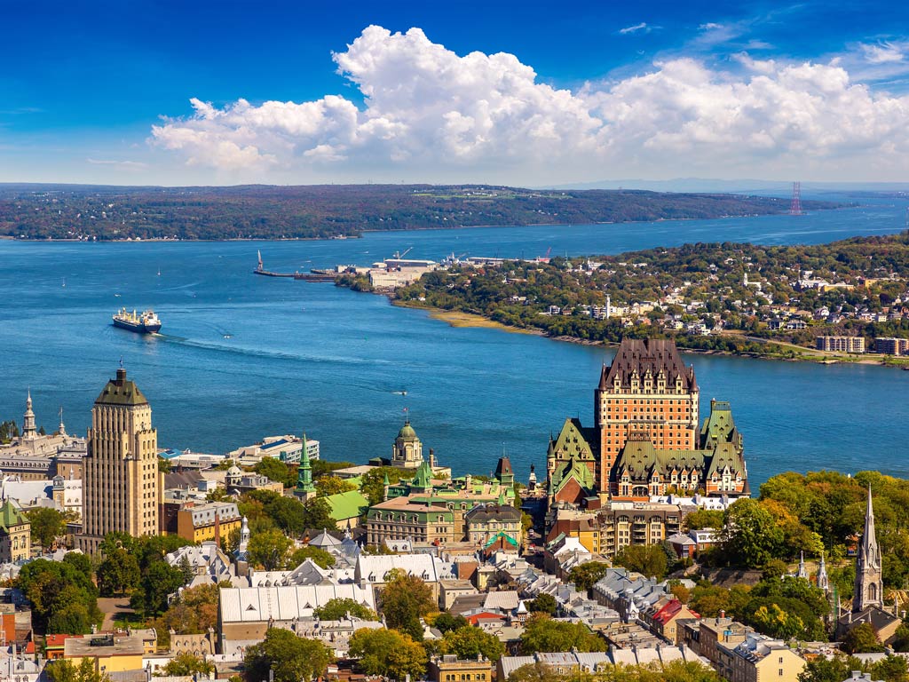 An aerial view of Quebec City, featuring the Château Frontenac, surrounding buildings, and the Saint Lawrence River—a beloved Canada fishing locale—under a partly cloudy sky.