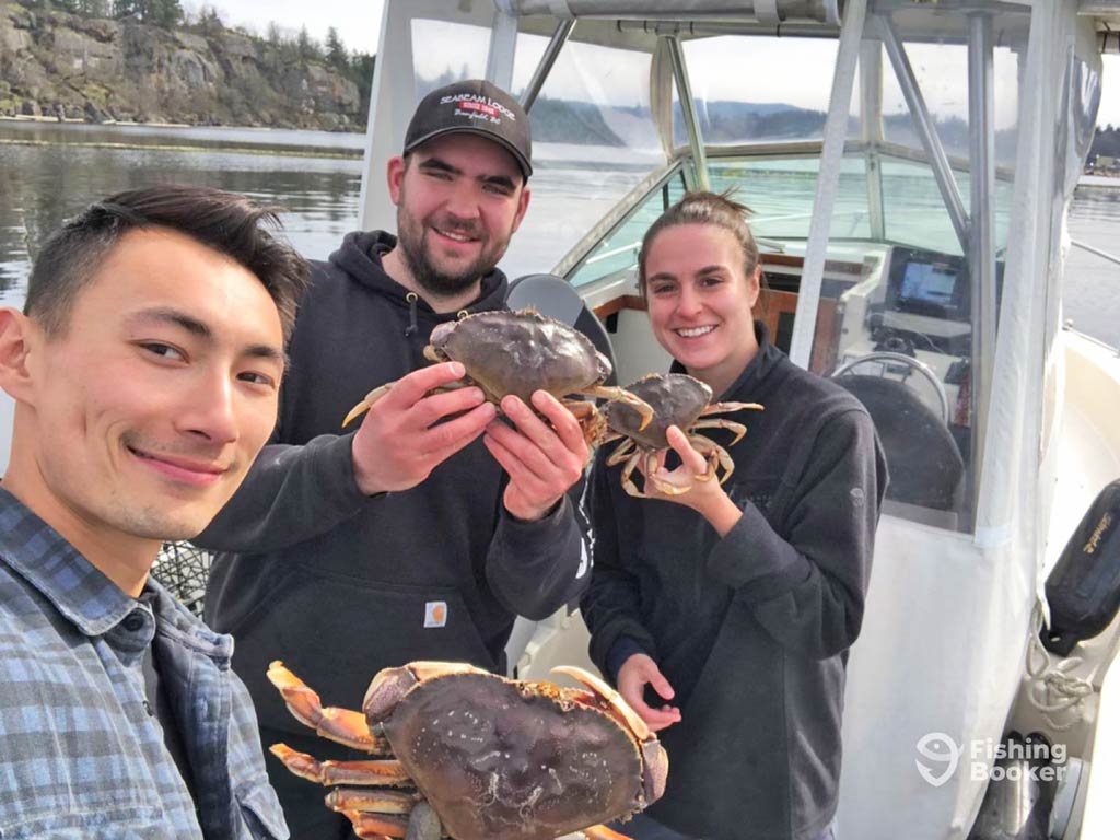 Three people on a boat in Canada are smiling at the camera while proudly holding their freshly caught Dungeness Crabs, enjoying a perfect day out on the water.
