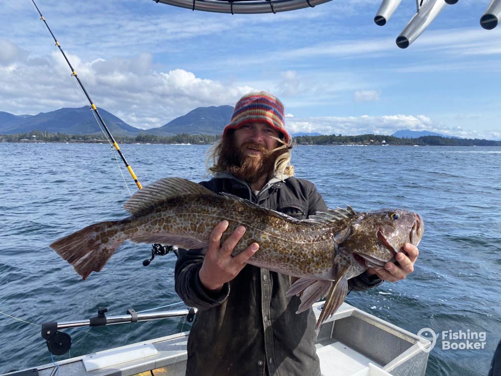 A person holds a large Lingcod with both hands on a charter, against the backdrop of mountains and water. They're wearing a colorful hat and dark clothing while on a Canadian fishing adventures.