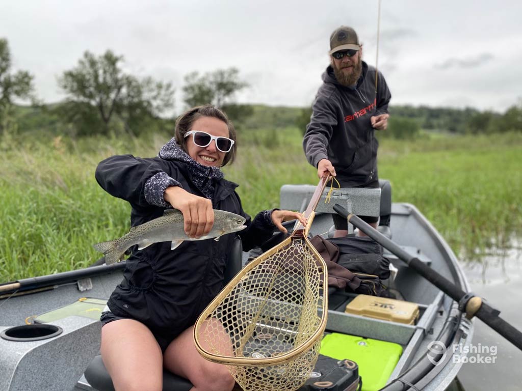 Two people are on a charter boat; one is holding a Trout, the other has a fishing net. They're enjoying Canada's rich fishing scene in Calgary, surrounded by grassy fields and overcast skies. 