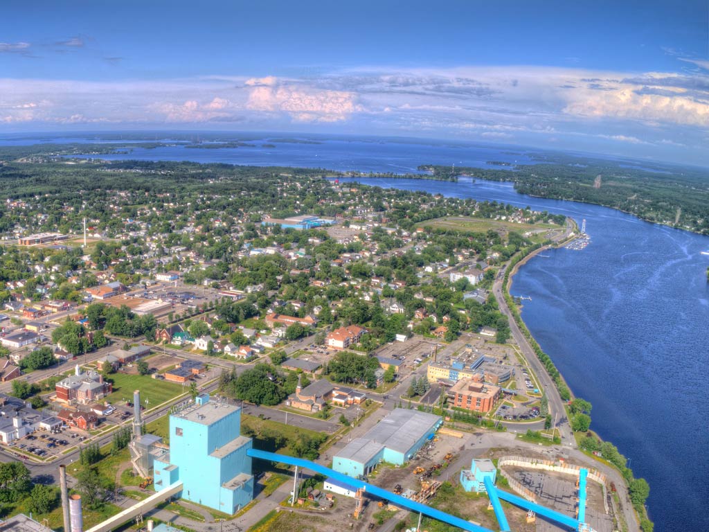An aerial view of Fort Frances with a river on the right, showcasing numerous buildings, roads, and greenery under a partly cloudy sky.