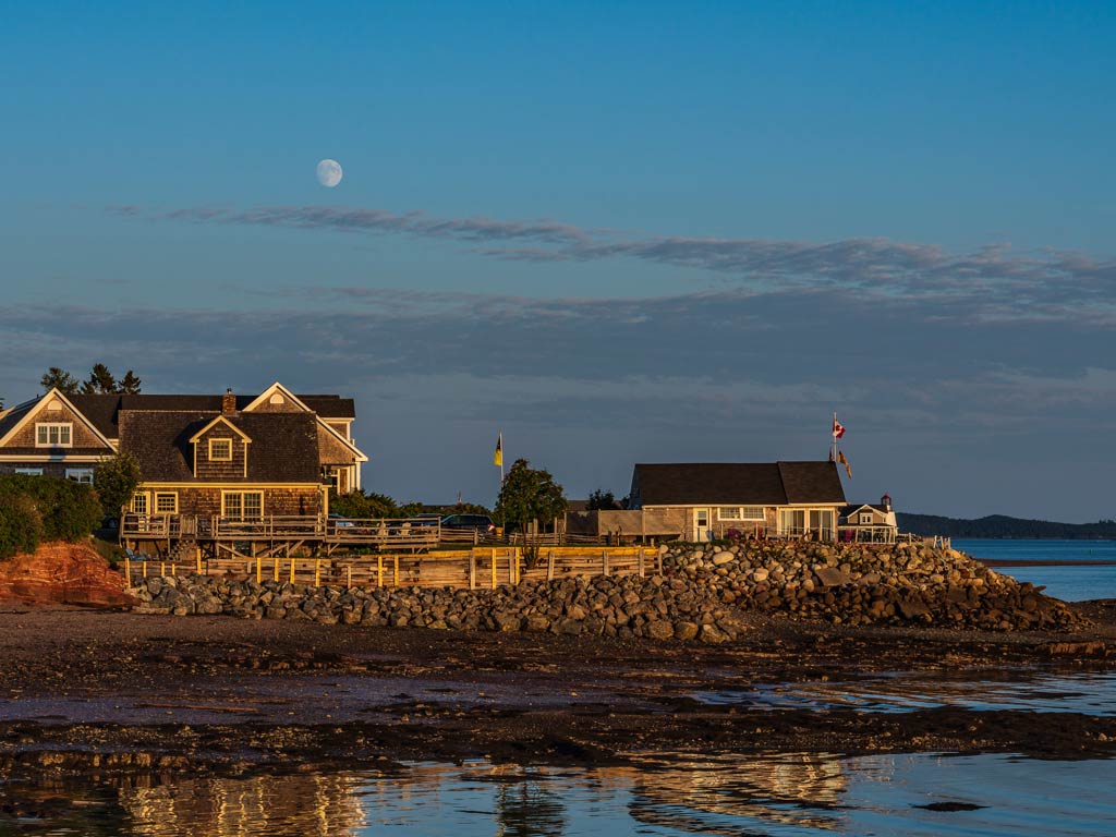 Coastal houses by the rocky shoreline in St. Andrews, with the ocean and a visible moon casting a magical glow in the early evening sky.