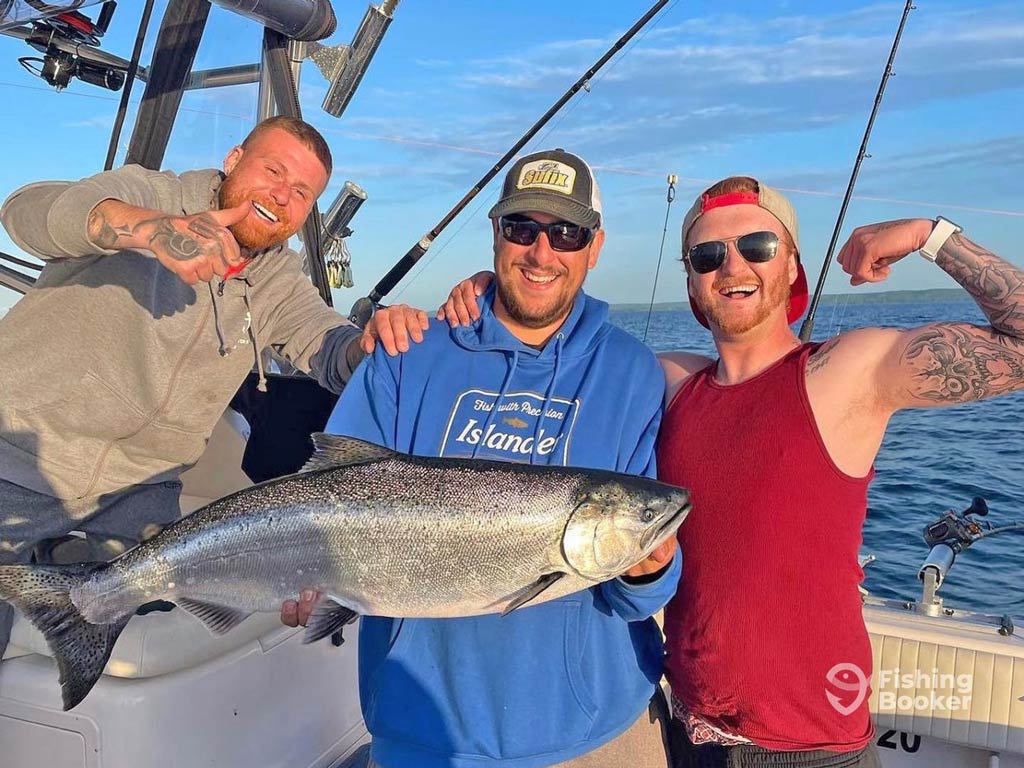 Three anglers are holding a large Salmon; one anglers’s pointing, another's wearing sunglasses, and the third's flexing his arm with the ocean in the background.