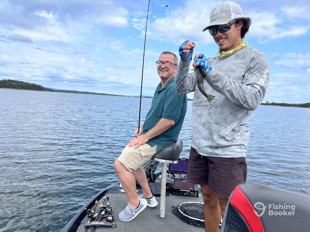 Two men enjoy Lake Hamilton fishing; one is sitting with a rod while the other is standing, smiling proudly with a small fish.