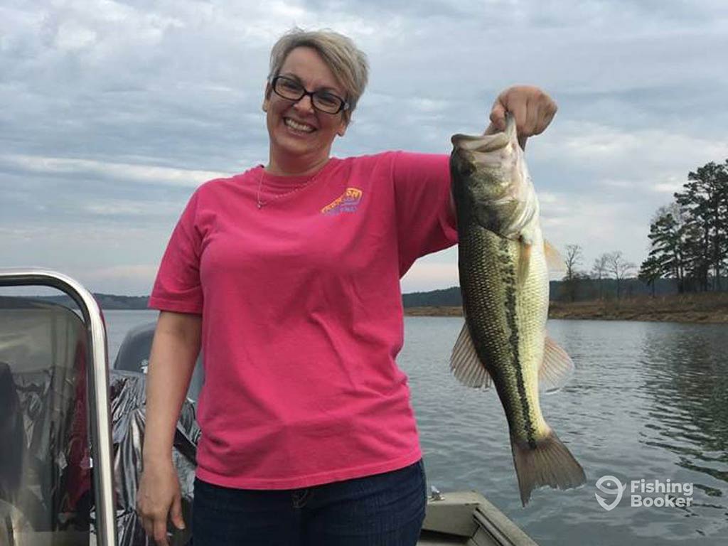 A woman on a boat is holding a Largemouth Bass and smiling. They're wearing a pink shirt and glasses on Lake Hamilton, with water and trees in the background.