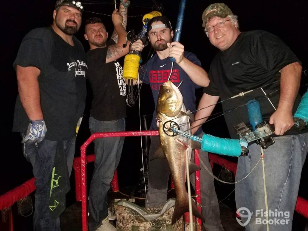Four men are on a boat under the night sky near Lake Hamilton, holding bowfishing gear and proudly showing off a large Catfish.