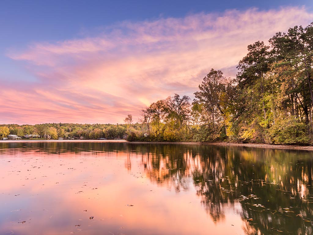A tranquil Lake Hamilton at sunset features a sky with pink and purple hues. Autumn foliage lines the shoreline, reflecting in the water, while fishing spots dot this picturesque scene.