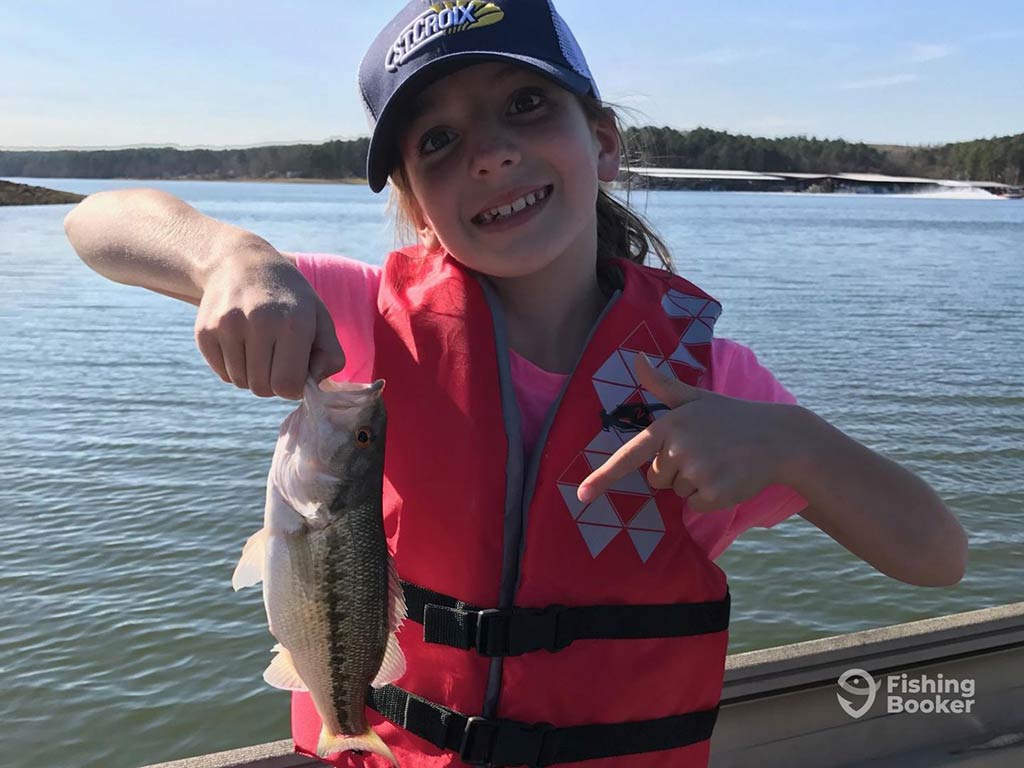 A child in a life jacket and cap proudly displays their Spotted Bass catch on a boat in Tennessee on a clear day.