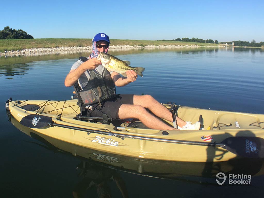 A person in a kayak holding a Bass on a calm lake with a clear sky.