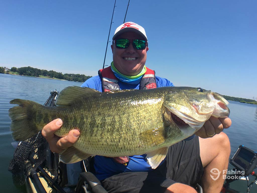 A person in a kayak holding a large Largemouth Bass, wearing sunglasses, a blue shirt, and a cap. There's a beautiful lake and trees in the background.