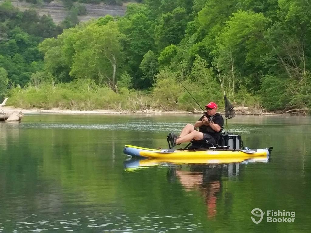 A person in a red cap fishing on a yellow kayak in a calm river surrounded by lush green trees.