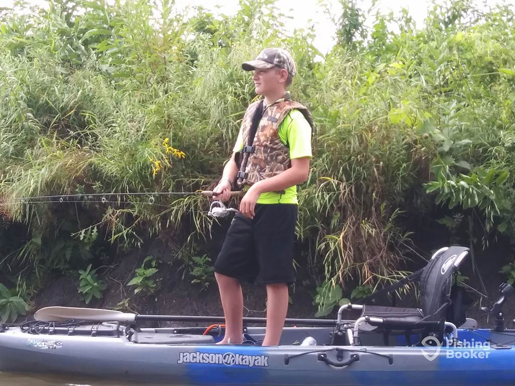 A young person's standing on a kayak, wearing a camouflage cap and vest, holding a fishing rod while surrounded by green foliage and water.