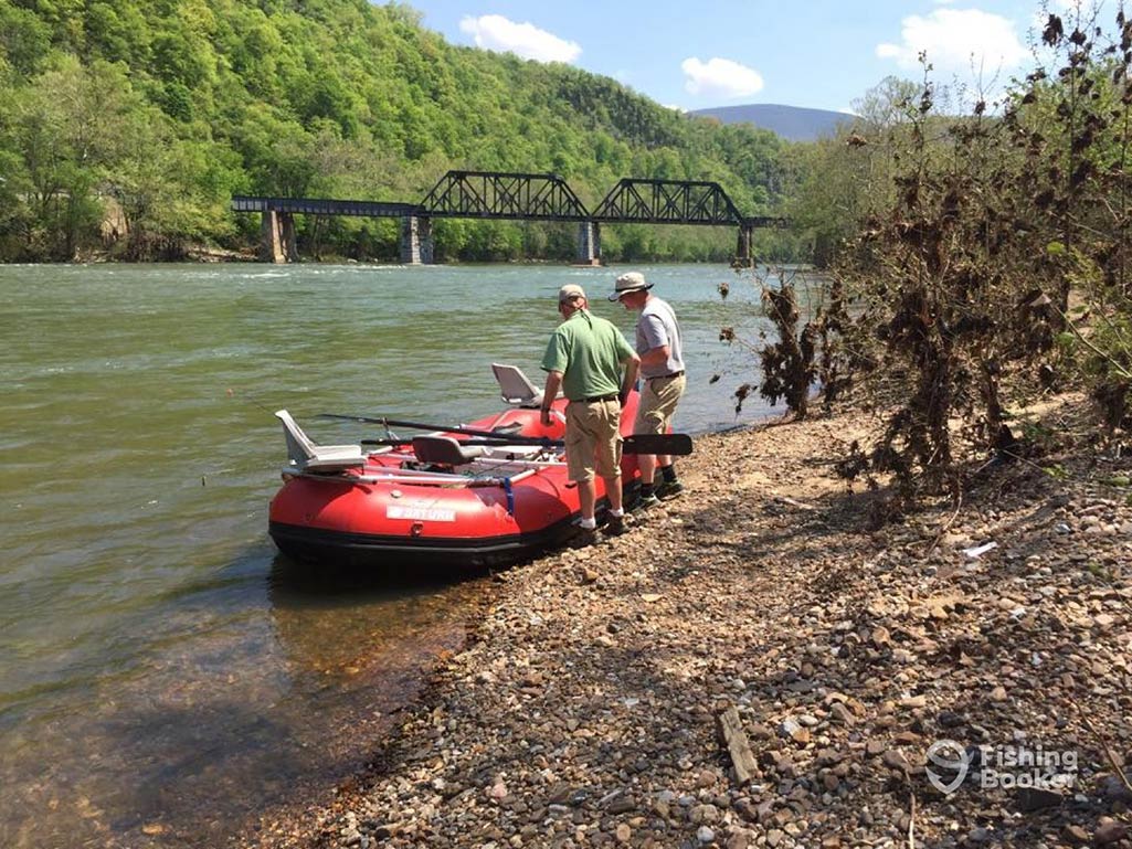 Two people are standing next to a red inflatable raft on a rocky riverbank, with a bridge in the background and greenery all around.