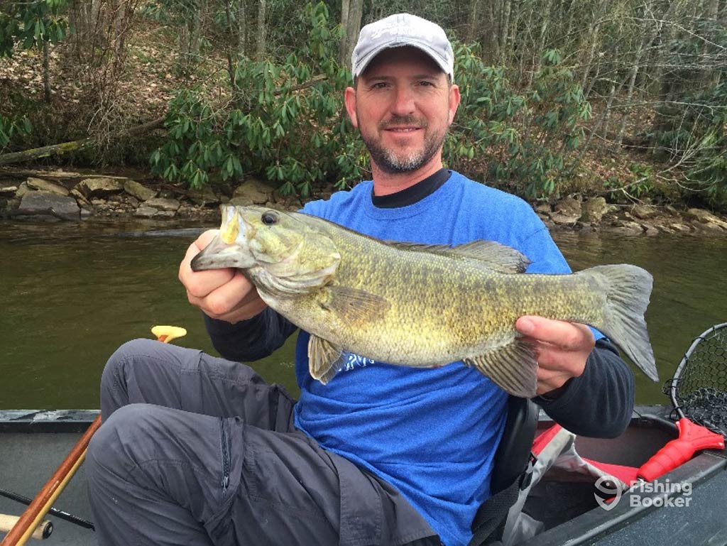A person sitting in a boat is holding a large Fish, wearing a blue t-shirt and gray cap. You can see a paddle, fishing net, and trees in the background.