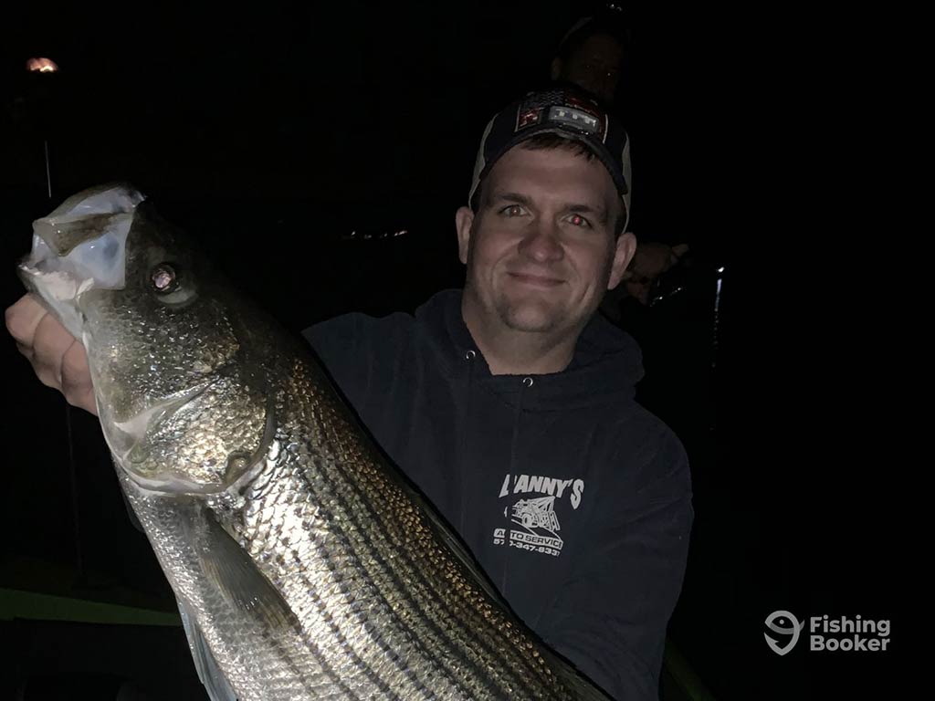 A man smiling while holding a large Bass fish, wearing a dark hoodie and cap against a night backdrop.
