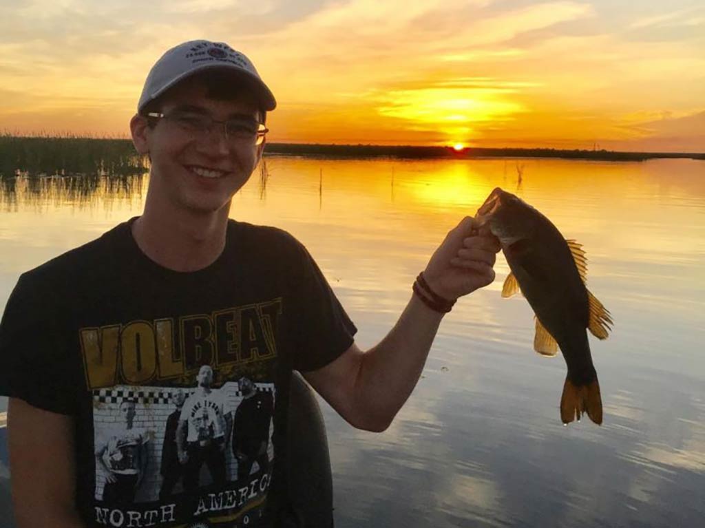 A person proudly holds a Bass fish at sunset next to a lake, wearing a cap and band t-shirt.