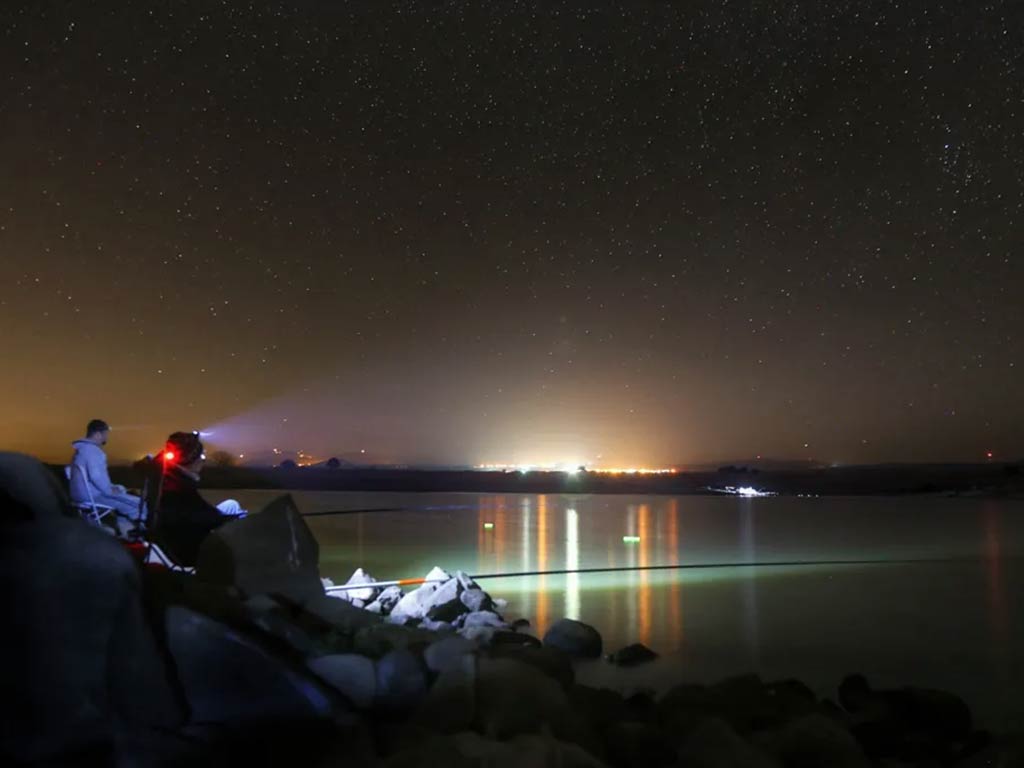 Two people are sittiing on rocks by a calm lake, Bass fishing at night under city lights and a starry sky. A flashlight beam is visible from a headlamp.