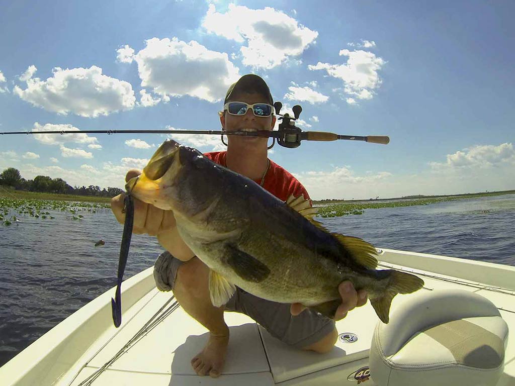 A man sitting on the bow of a boat and holding a Largemouth Bass with a soft plastic lure dangling from its mouth, as he holds his fishing rod between his teeth