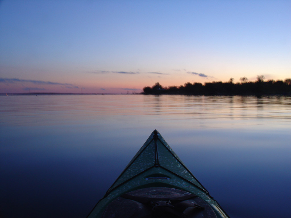 A photo of Devils Lake at sunset, with clear skies and trees silhouetted on the horizon. The serene setting is gently decorated with dancing reflections on the water's surface.