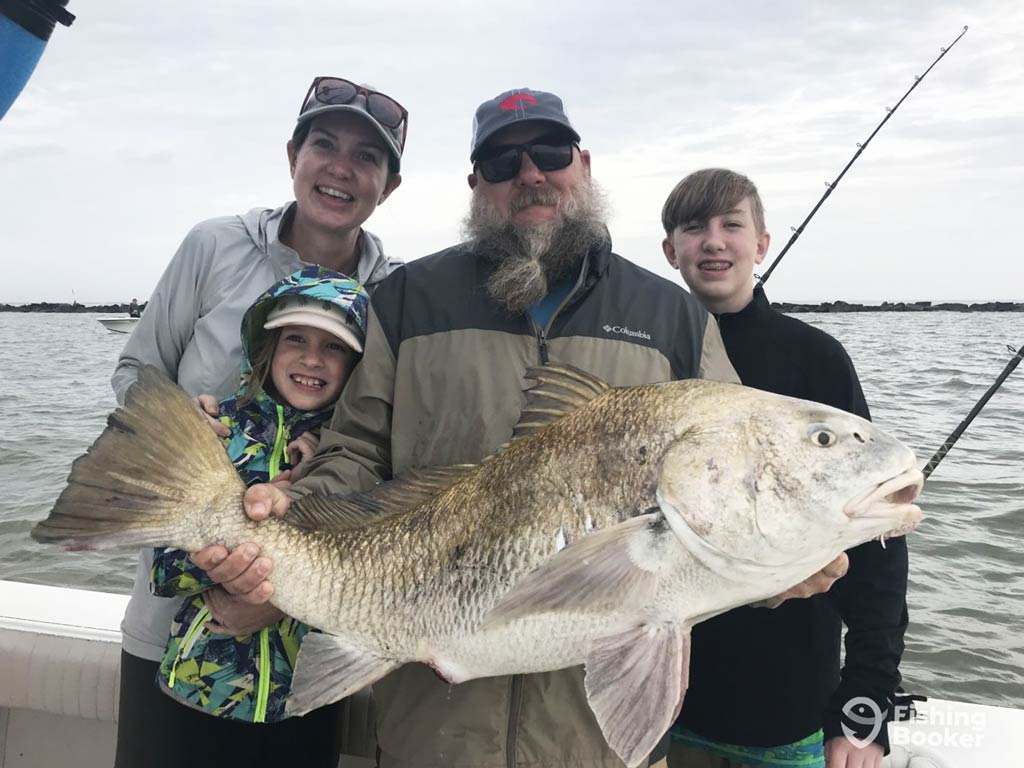 A group of four people is smiling on a boat, holding a large fish they caught.