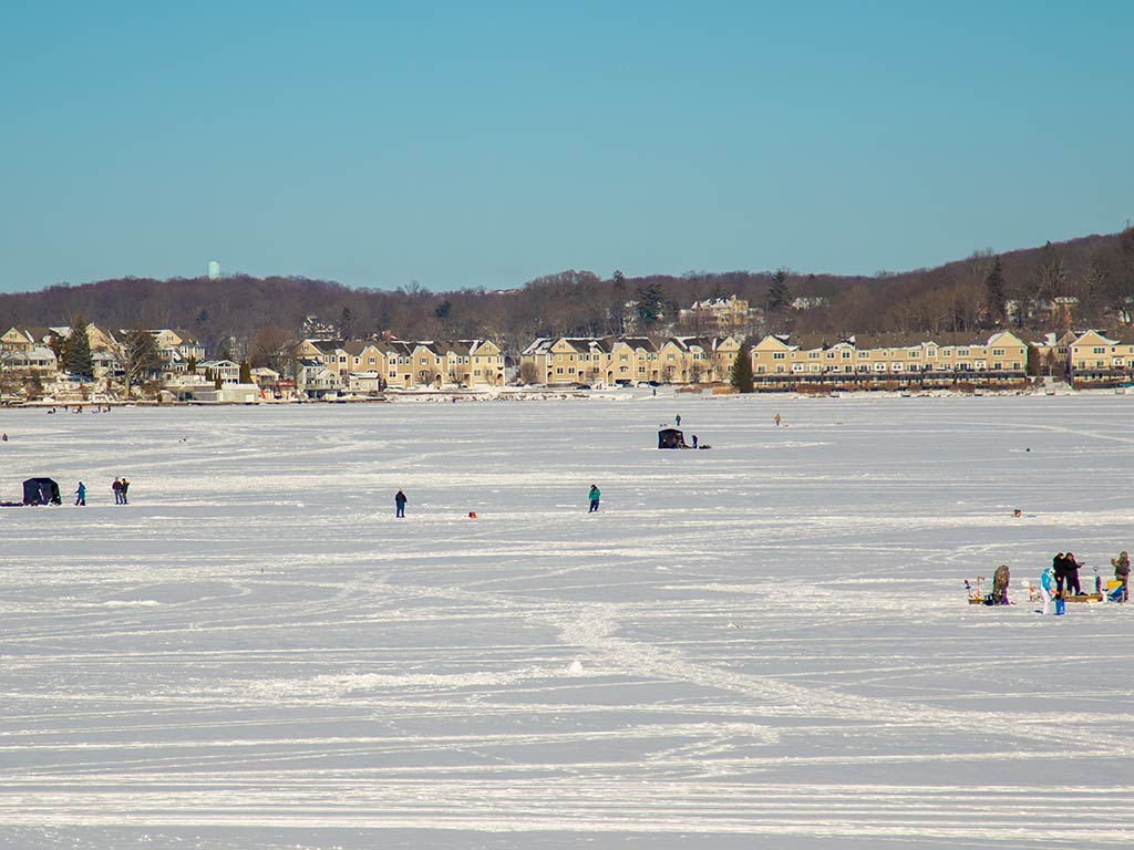 A view across Lake Hopatcong as it's frozen over and covered with snow, with a number of ice huts and ice fishers on the lake on a clear day