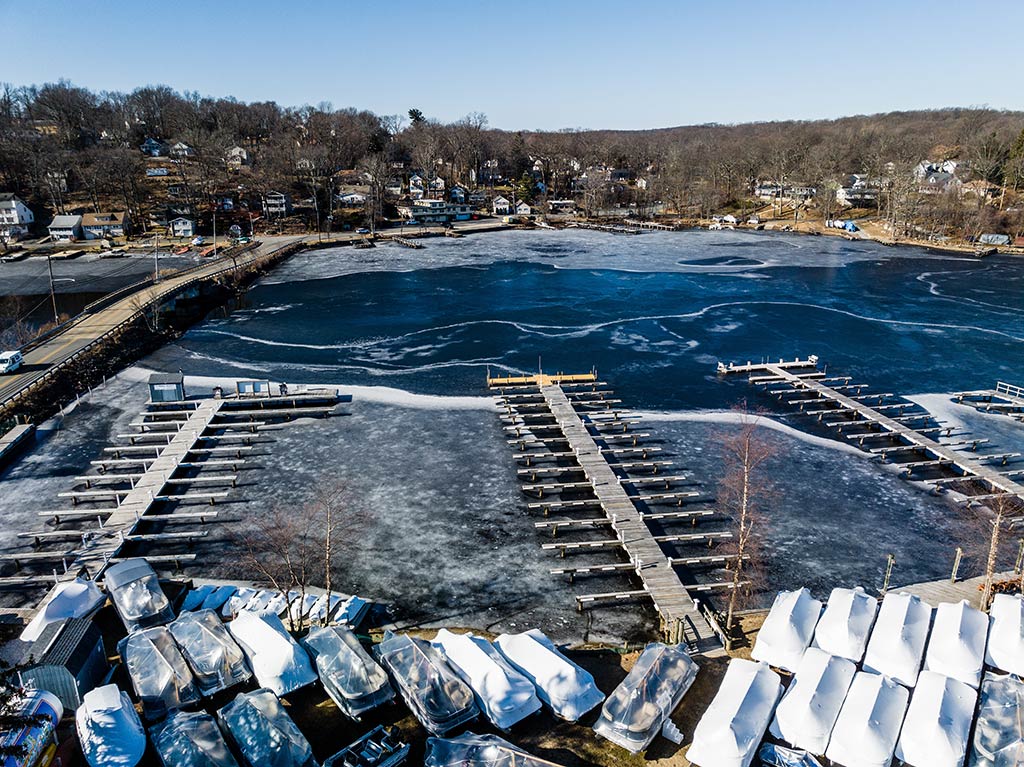 An aerial view of a frozen marina on Lake Hopatcong on a cold winter's day with numerous covered boats at the bottom of the image and a town visible in the distance