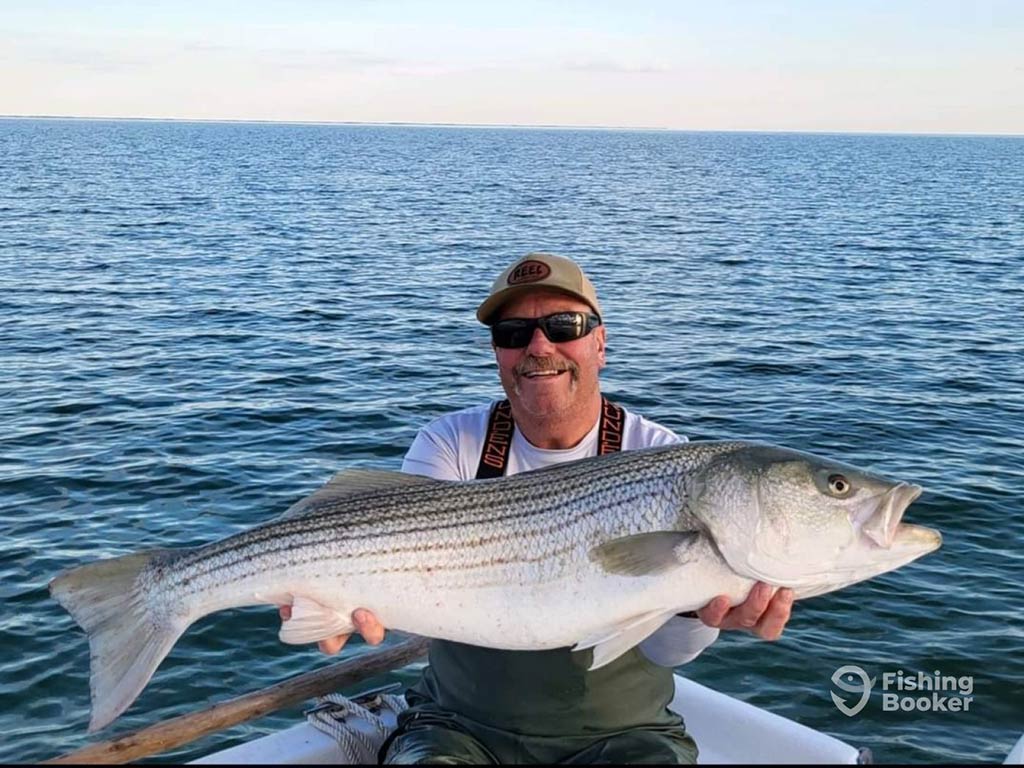 A man in sunglasses and a cap proudly holding a large Striped Bass with open waters in the background.