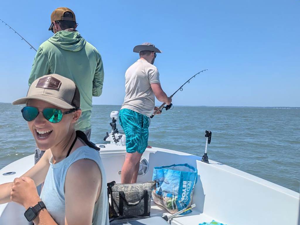 Three people are fishing on a boat near Assateague Island under a clear sky. A woman in sunglasses and a cap is smiling in the foreground, enjoying the serene day. They're hoping to catch some Striped Bass, Red Drum, or Flounder during their trip. There's a gentle breeze keeping them cool as they share stories and laughter while waiting for a bite.