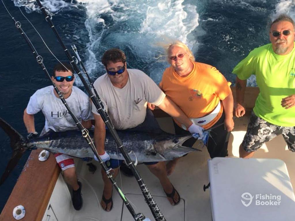 A view from a flybridge of a boat looking down at four people near Assateague Island holding a large Billfish, with fishing rods close by, celebrating their successful fishing adventure.