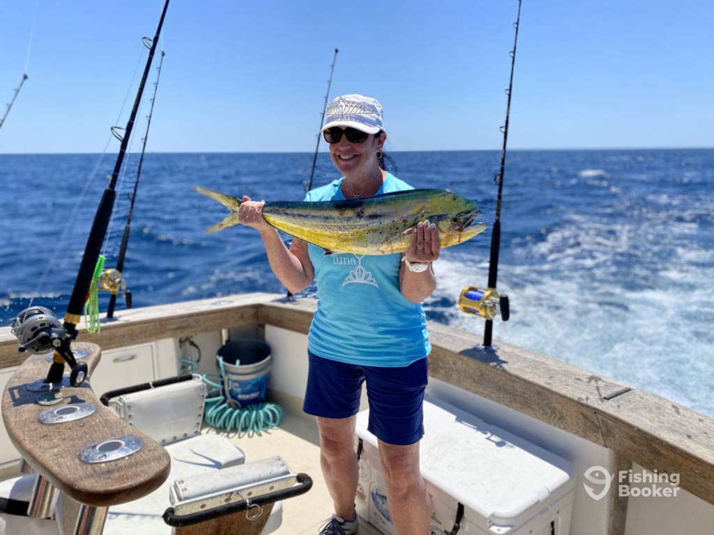 A woman proudly holds a Mahi Mahi on a boat, surrounded by multiple fishing rods, with the vast ocean in the background.