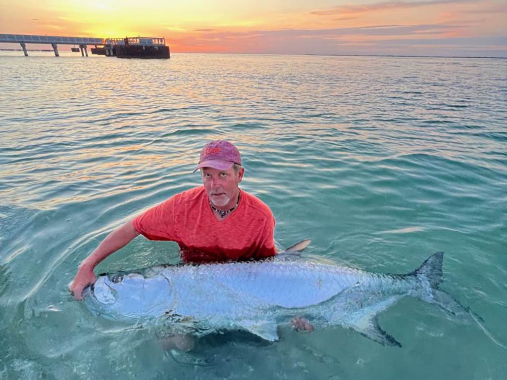 A man in a red shirt is holding a large Tarpon in shallow water at sunset near a pier.