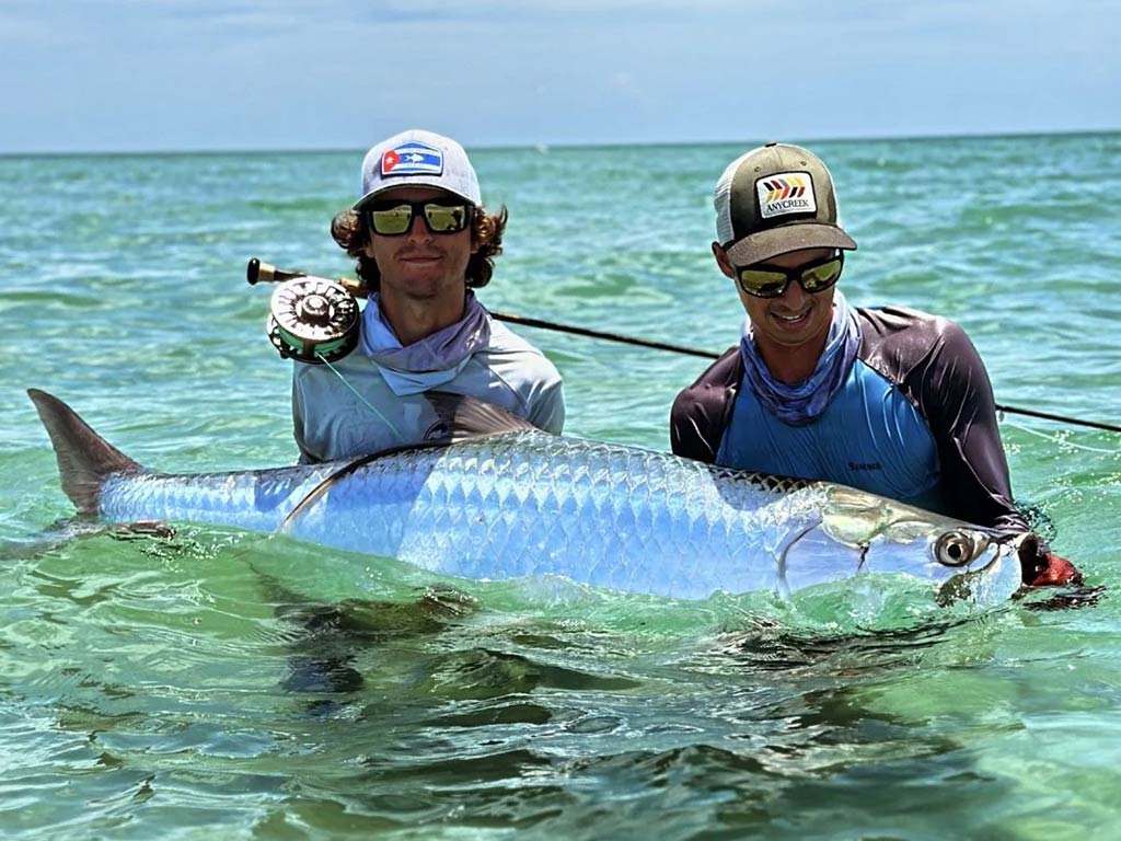Two men are standing in shallow water, each holding a large Tarpon. They're wearing hats, sunglasses, and fishing gear.