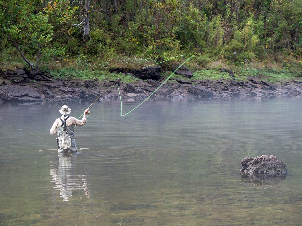A person stands in shallow water, fly fishing amidst trees and rocks.