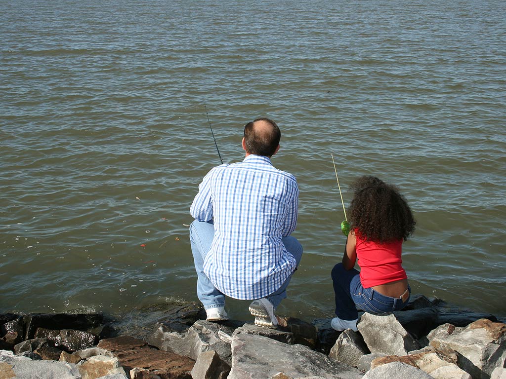 A view from behind of a man and child sitting on rocks by the water, their rods ready as they face a calm lake.