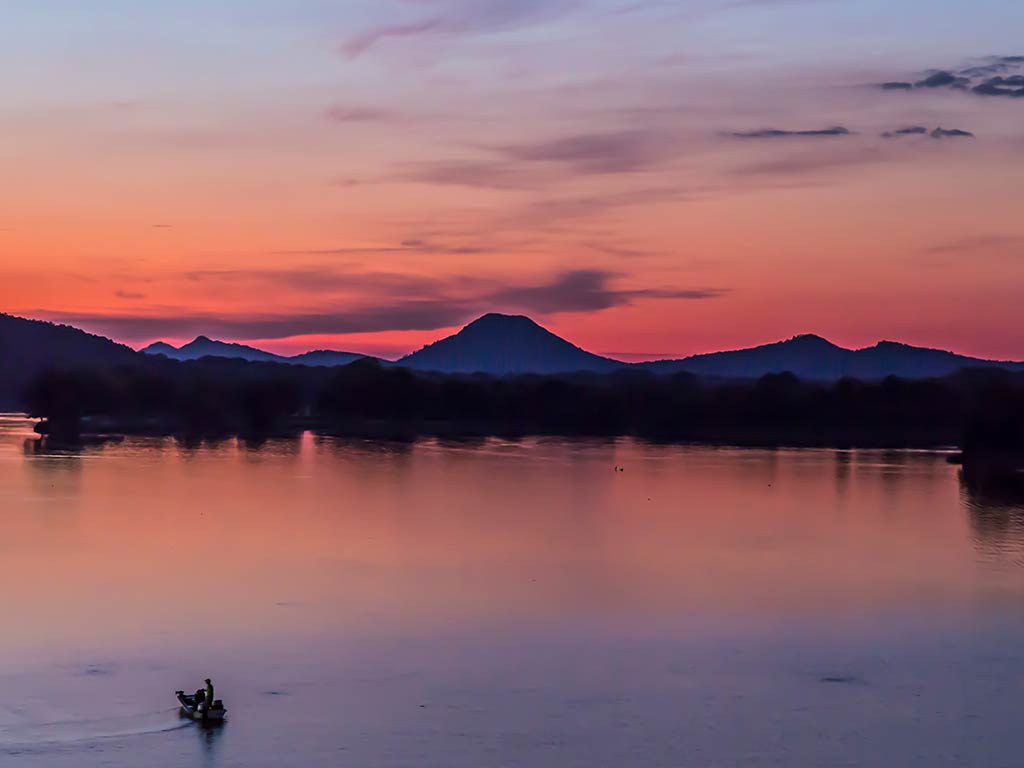 A small boat on a calm lake at sunset, with silhouettes of distant hills and a vibrant pink and orange sky.