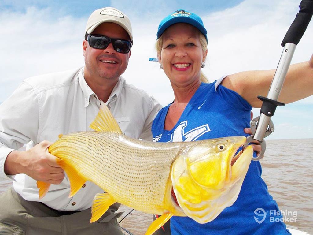 Two people are smiling while holding a large Golden Dorado with a fishing tool, standing on a boat under a cloudy sky.