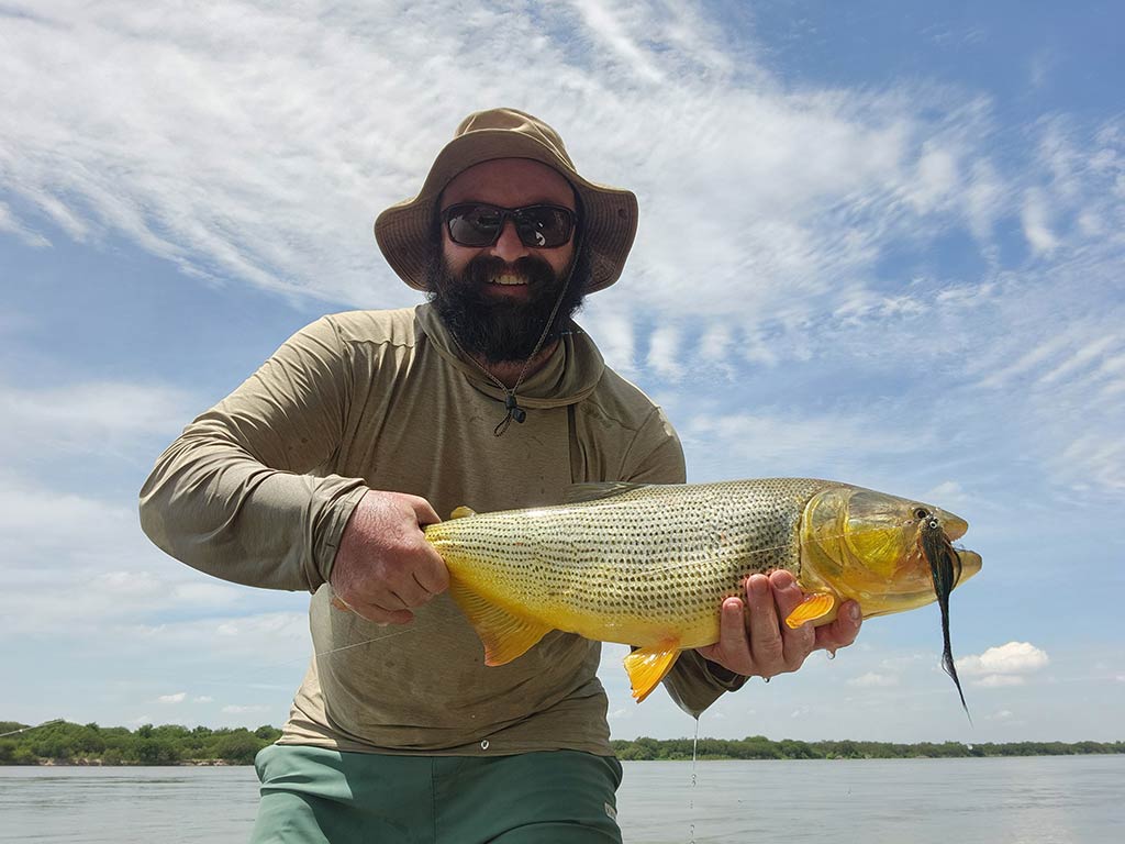 Someone proudly holding a Golden Dorado, its yellowish hue and stripes catching the eye. They're wearing a hat and sunglasses, set against a backdrop of bright blue sky and shimmering water.