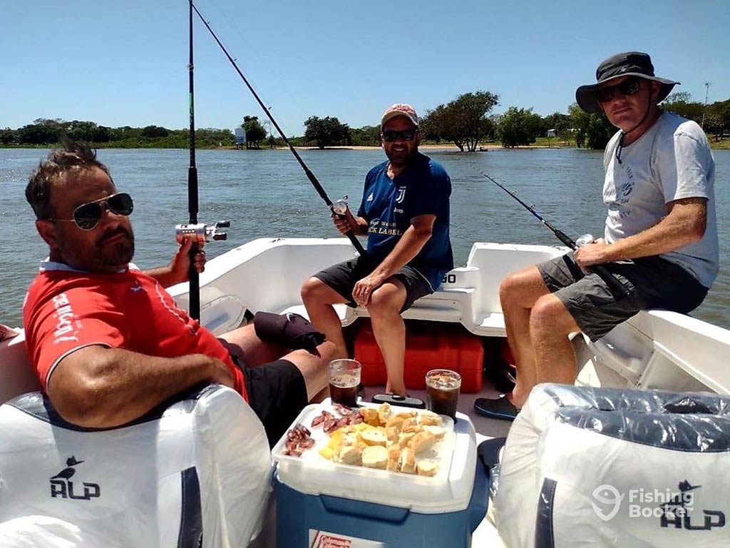 Three men sit in a boat, fishing under the sun, with snacks and drinks on a cooler in front of them, enjoying the perfect day.