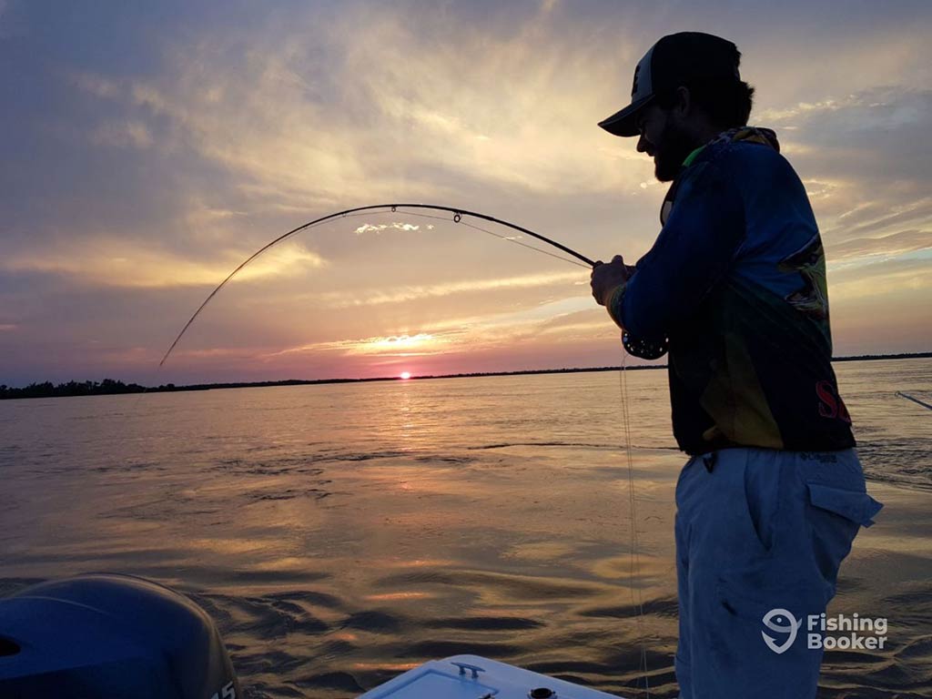 As the sun sets, someone is fishing for on a boat, their rod bending amidst a colorful sky and calm water, with the motor silently humming.