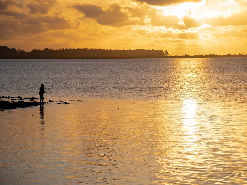 A silhouette of a person engrossed in fishing by the Uruguay River at sunset, with the golden light shimmering on the water.