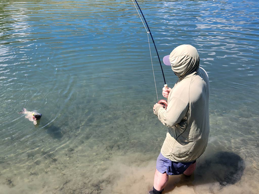 A person clad in a hood expertly uses one of the best freshwater fishing rods, reeling in a Fish from the clear blue waters near the shore.
