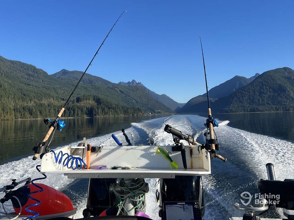 A boat with fishing rods and Salmon fishing lures cuts through the lake, surrounded by mountains, leaving a wake behind under the clear blue sky.