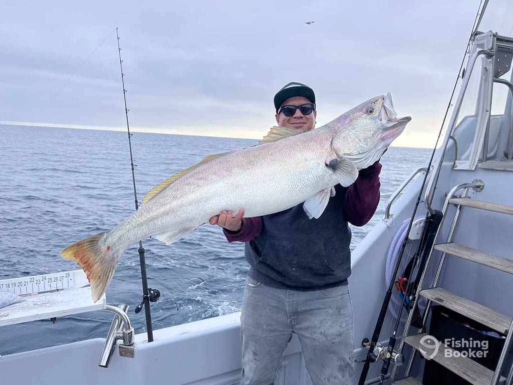 A person on a boat holding a large fish with both hands. Another fishing rod is visible nearby, while the ocean is in the background under a cloudy sky.