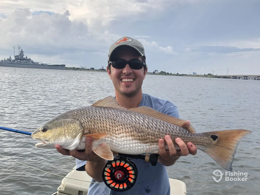A man in a baseball cap is smiling while holding a large Redfish on a boat. In the background, there's water and a battleship.