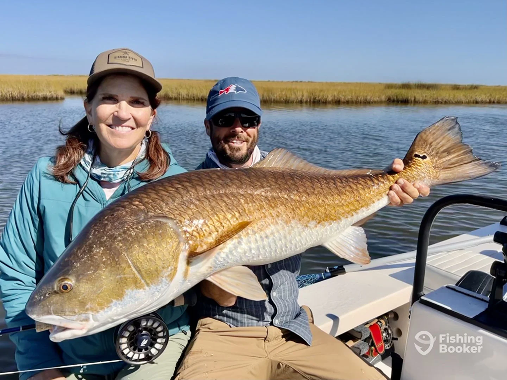 Two people are on a boat, holding a large Redfish in a marshy area. They’re both wearing hats and sunglasses on a sunny day.