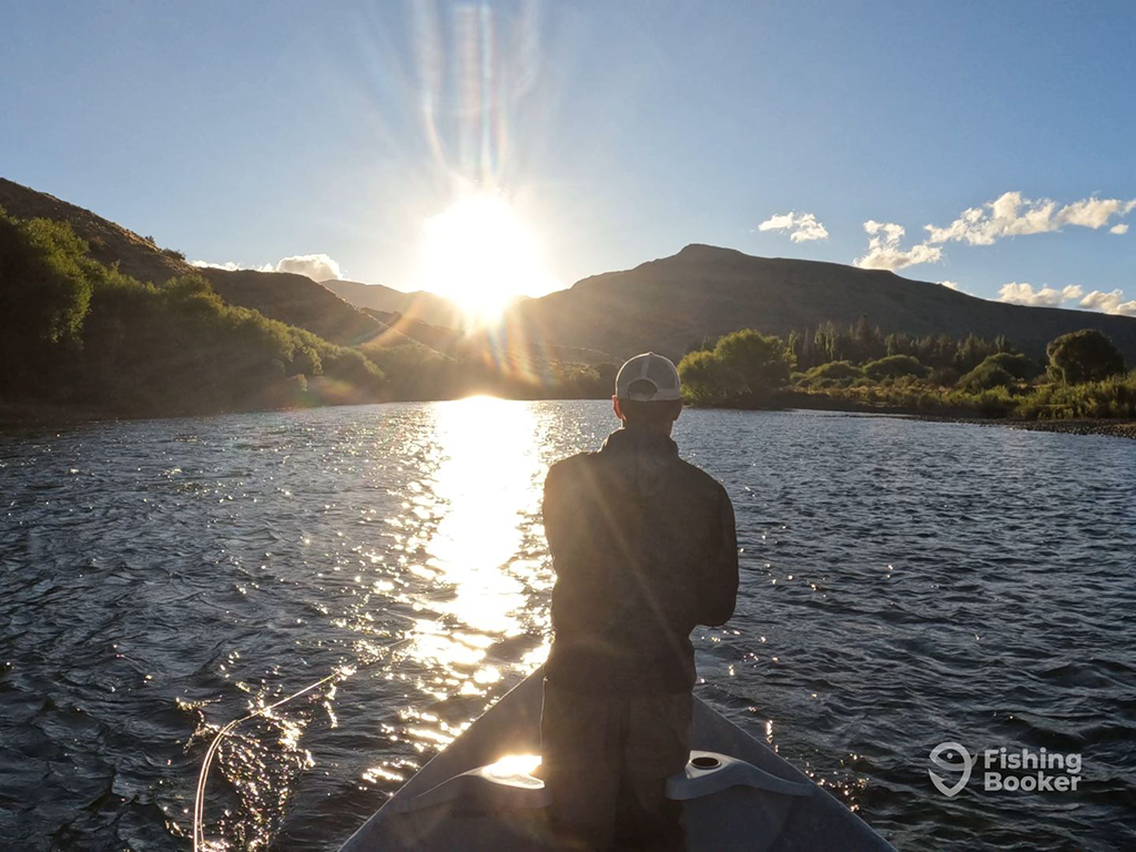 A person enjoying Trout fishing from a boat on a serene Patagonian river, with the sunrise casting reflections on the water and hills in the background.