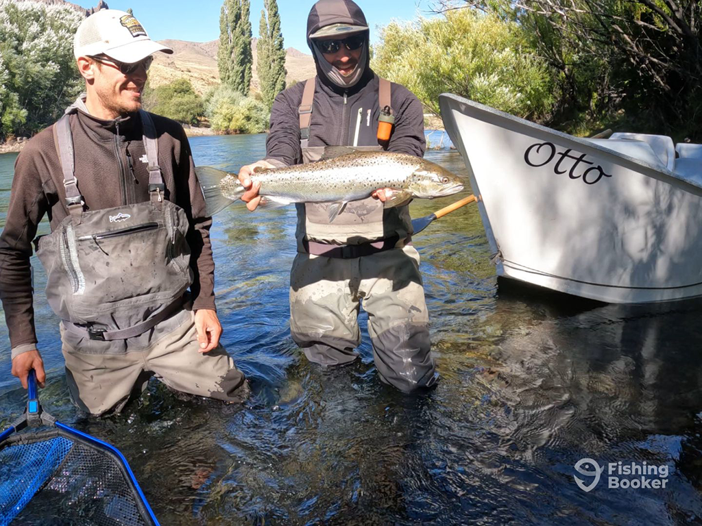 Two people are standing in a river, holding a large Trout caught while fishing in Patagonia. A boat named "Otto" is partially visible nearby.