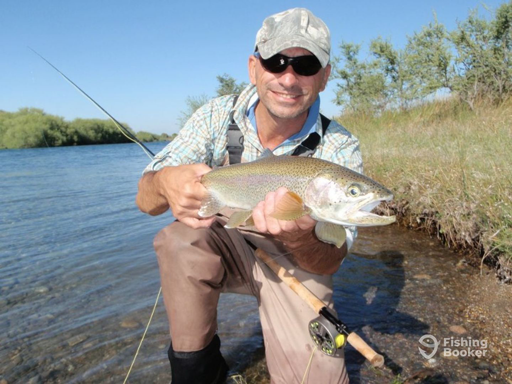 A man in a cap and sunglasses holds a large Trout by the riverbank, standing proudly in chest waders with his fishing rod. He's showcasing the thrill of Trout fishing in Patagonia.
