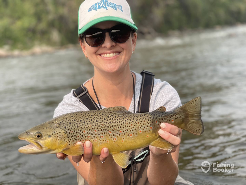 A woman wearing a hat and sunglasses is holding a Brown Trout by a river, smiling at the camera.