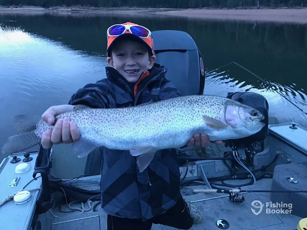 A child in a cap and jacket is holding a large fish on a boat, smiling. A calm body of water's visible in the background, with trees lining the shore.